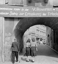 Drei Menschen gehen im August 1950 durch das mit einem Banner für die ersten DDR-weiten, sogenannten Volkswahlen und Friedensparole versehene Klostertor in Kamenz in Sachsen